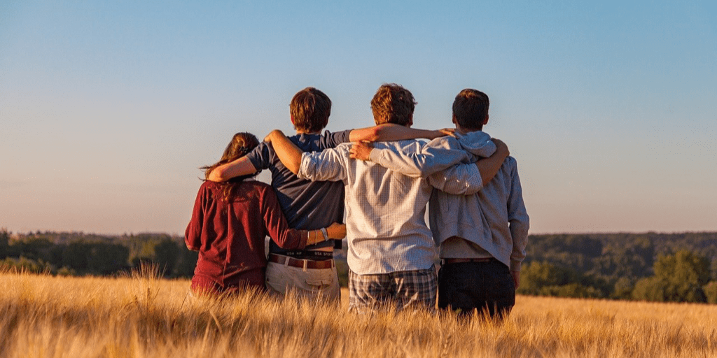 A group of four teenagers seen from behind holding their arms around one another in a natural setting