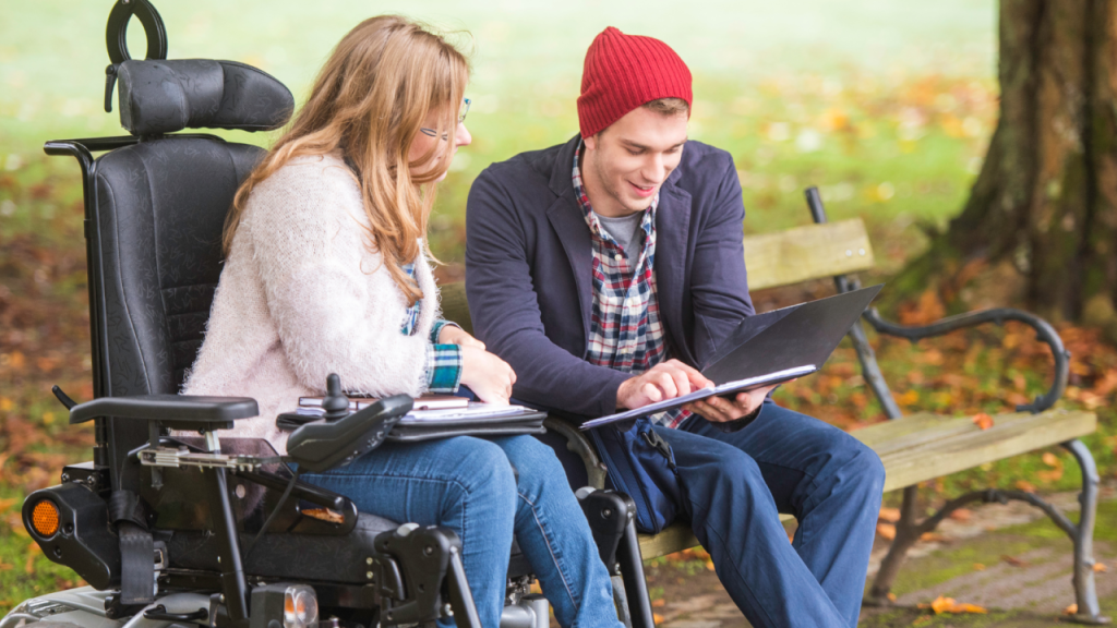 A person on a wheelchair in the park with a young person sitting on a bench next to them and showing them a book