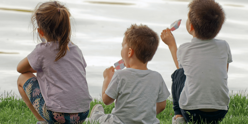 Three children seen from behind as they eat ice lollies in front of a lake