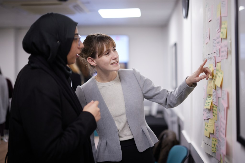 Two people looking and pointing at a board covered in post it notes.